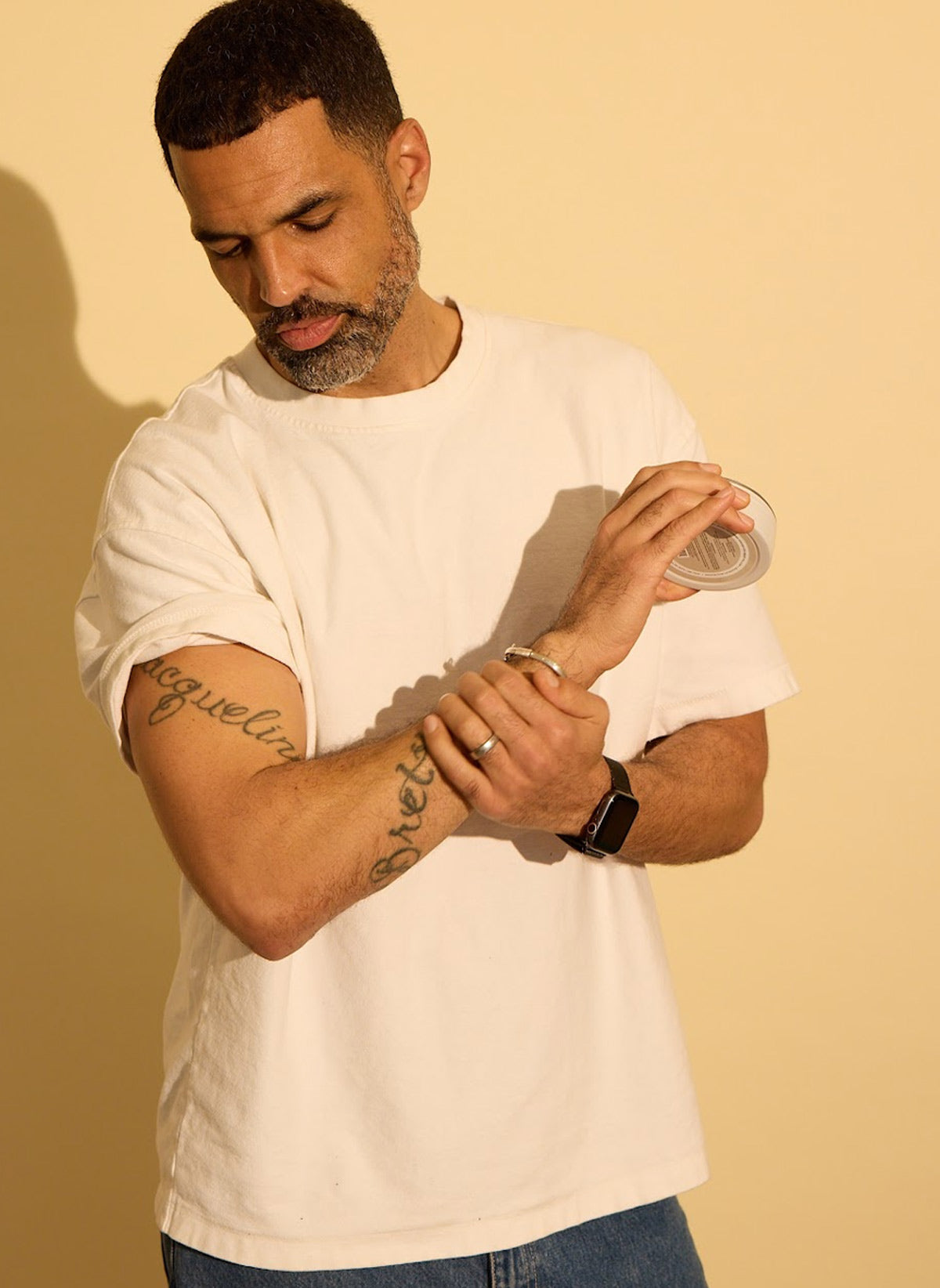 Man in a white t-shirt holding a grass-fed beef tallow face cream in tin jar against a beige background